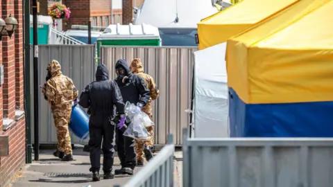 Getty Images Four people in protective overalls and masks walk into a building in Salisbury. The area is heavily cordoned behind metal fences and there are a number of large forensic tents in the street. 