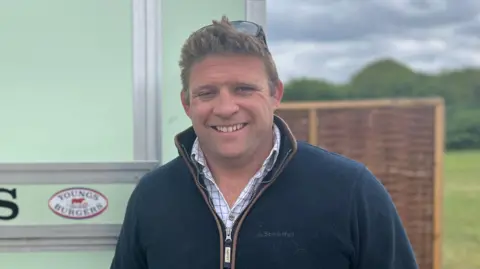 A man wearing a blue zip-up fleece and white checked shirt smiles at the camera. He has blond hair with sunglasses perched on top. 