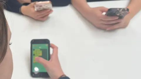 A picture of schoolchildren on their mobile phones, sat surrounding a white table.