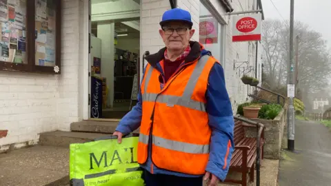 A man in his 70s wearing glasses, a blue cap and blue jacket with a flourescent orange high-vis vest. In his right hand he is holding his flourescent yellow paperbag with 'Mail' printed on it in capital letters. He is standing outside a whitewashed post office.