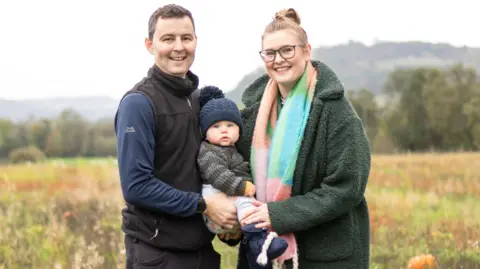 Family Picture On the left there's a man with dark hair and eyes. He's smiling at the camera, showing his teeth. He's wearing a black gillet with a dark blue long sleeve top underneath. He's holding a baby which has blue eyes and is looking at the camera. The baby hhas a navy bobble hat on his head and is wearing a stripped knitted green and grey jumper. To the right is a woman with light brown hair. She's also smiling at the camera. She's wearing glasses a green coat and a blue, green, pink and orange scarf.