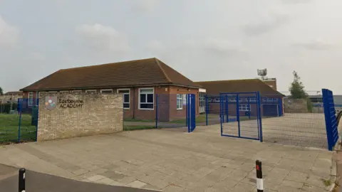 Google A yellow brick wall with the logo of The Eastbourne Academy in front of red brick, single-storey school buildings with sloped roofs. The school buildings are surrounded with a blue wire fence.