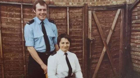 Thames Valley Police Ed and Madge, in police uniform. Ed is standing behind Madge, who is sitting down. He has on a blue skirt, with a black tie and black trousers, Madge, a white shirt, with a black tie, and is sitting down. They both have short dark hair.