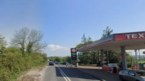 A rural two-lane road with hedgerows on each side, a Texaco garage sits on the right hand side of the image. 