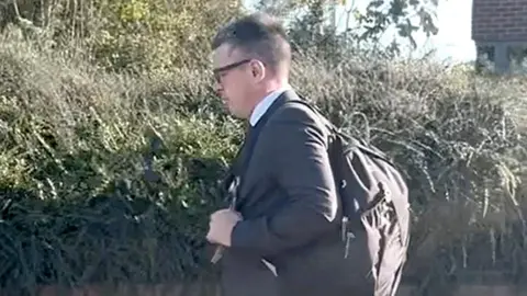 PA Media A man in a dark suit and carrying a back pack walks on a street, a hedge is visible behind him