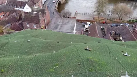 A large area of green netting with metal spikes sticking out of it and in the distance there are a number of houses, a road and a bridge over a river.