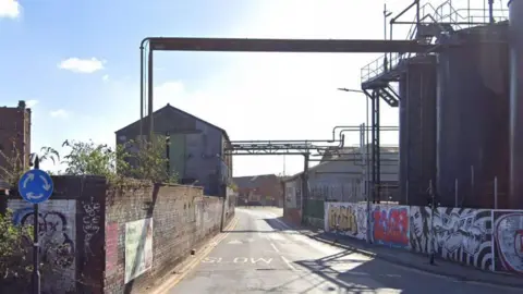 Google Two industrial buildings stand at either side of a road. Pipework stretching over the carriageway, between the two buildings, is visible. On the right side, two large black silos stand behind a wall covered in colourful graffiti.