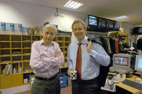 Humphrys - here with former Prime Minster Tony Blair. He crosses his arms, wearing a pink shirt and jeans with a belt. Blair wears a light blue shirt with bronze tie, and holds his blazer over his shoulder. They are standing in an office, with computers in the background. 