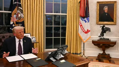 Jim Watson/Getty Images President Donald Trump, a man with short, blond hair, sitting at a wooden desk in the Oval Office speaking to the media. On the wall to his left there is a gold-framed painting of President Andrew Jackson. Trump is wearing a dark suit jacket, a white shirt and a purple tie. There are document folders on his desk. Behind him there are flags and large windows draped with gold-coloured curtains. 