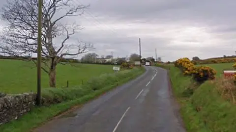 A section of the A26 Braaid Road. The tarmacked road has green fields on either side, which include a tree on the left and yellow flowering gorse buses on the right. 