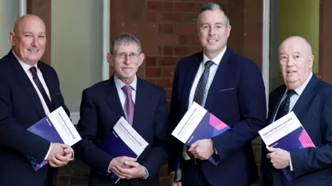 Department of Education Four men are standing together. All four are holding a document. They are all wearing navy suits with white shirts. 