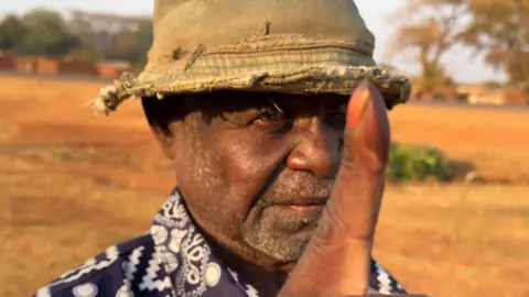 A head shot of a man, wearing a battered, khaki, cloth hat