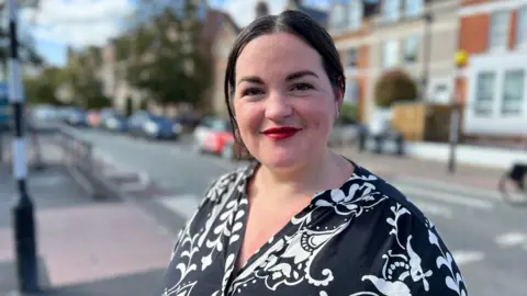 A smiling Heather Williams looks at the camera. She wears a black top with a white pattern. A blurred street is in the background 