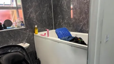 Charlie Stubbs / BBC Through a white doorway, there is a high white bath with a blue chair inside. On the bath's rim, there are three cleaning bottles. Next to it is an open window with a small circular mirror and bottles next to it. There is also a small sink and the back of a wheelchair.
