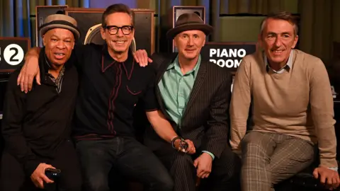 The image shows four men sat smiling for a photo. They are all wearing long sleeves and two are wearing hats.