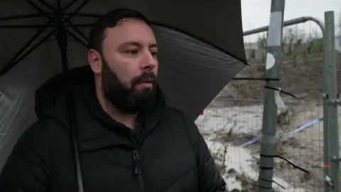 James stands in front of a fence, rubble can be seen behind it. He wears a black coat and holds a black umbrella. He has short dark hair and a dark beard