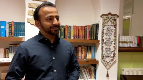 Manik is looking at the interviewer to the side of the camera and smiling. He is wearing a dark blue shirt with white dots on. Behind him are religious text books on shelves.