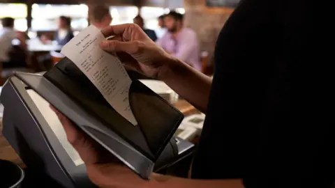 A waitress dressed in a black shirt holds a bill in a black envelope. 
She is standing near to a till in a restaurant. 
A number of people can be seen sitting at tables in the background. 