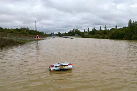 Joe Giddens/PA Media The open boot of a car is visible above the water where the vehicle is submerged in flood water on the A421 in Marston Moretaine, Bedfordshire.