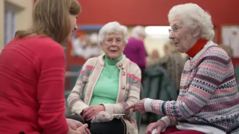 'I received a bank card after dropping the winter gasoline fee' A group of three women sat in a circle. Younger woman wearing a red jumper chatting to two older women wearing knitted jumpers. One of the women is holding some knitting
