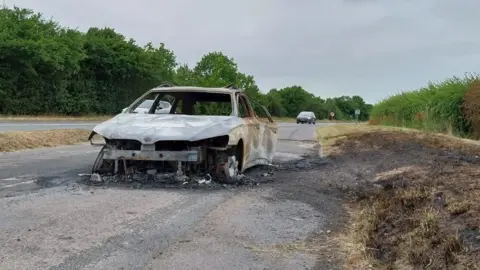 Owen Ward/BBC A white car is sitting on a slip road off of a main road. The car had been on fire which has resulted in fire damage, taking off the front off the car and the windows.