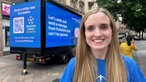 A woman with long blond hair wears a blue charity T-shirt and smiles as she stands next to a blue van displaying digital billboards for Yorkshire Cancer Research. Messages read: "Send a white rose to London", and "Let's unite and send a message from Yorkshire that can't be ignored". 