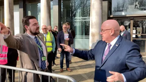 Two men in suits appear to be rowing behind a metal barrier