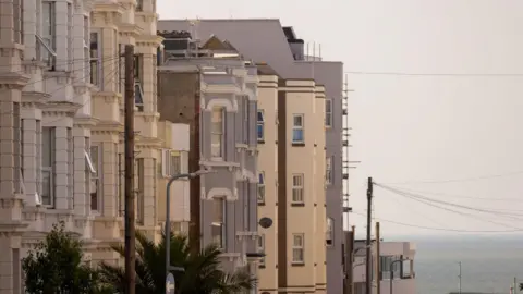 A row of light-coloured terraced buildings on a clear day. The sea can be seen in the background.
