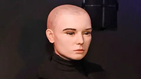 PA Media Uma estátua de cera da cantora Sinéad O'Connor vestindo um suéter preto e um casaco preto