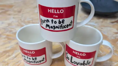 A trio of irrevent message mugs made at 1882 Ltd, white with handles, decorated with red label design, saying 'Hello, I'm born to be Magnificent'
