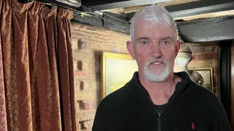 A man with white hair and a white beard is wearing a black fleece and standing in what looks like an old house at night time, lit by a lamp not seen in shot. There is a suit of armour behind him.