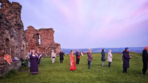 Charlotte Knaggs 12 people stand facing the rising sun at Sandal Castle - a medieval ruined building. 