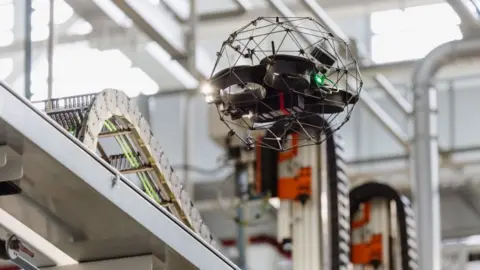 JLR A drone flies around the warehouse by some shelving with equipment on. It is black with a circular metal framework arounds what could be a camera.