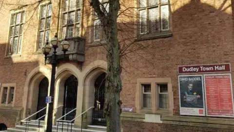 Google The entrance to Dudley Town Hall with steps and railings leading up to the arched doors. There is a sign that reads Dudley Town Hall at the front of the brick building and there is a tree with bare branches and a lamppost.