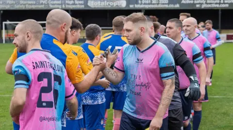 Angels United players shaking hands before a match against Sands United FC. The Angels' players are wearing pink and blue shirts, and navy shorts.