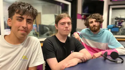 Jon Wright/BBC Kieran, Jack and Bailey sit inside a radio studio. One young man on the left is wearing a white t-shirt, in the centre, a young man wears a black t-shirt with long hair tied back, and far right is a man with long brown hair and a hooded top that is blue, white and pink.