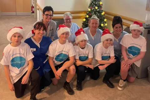 The Denny Boys wearing white T-shirts and santa hats while kneeling in front of a small Christmas tree with staff at Strathcarron Hospice.