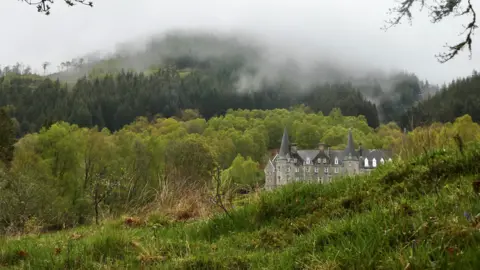 Ian Pitkethly Huge grey house with two pointy roofs nestled in green countrside beside a hill covered in trees.