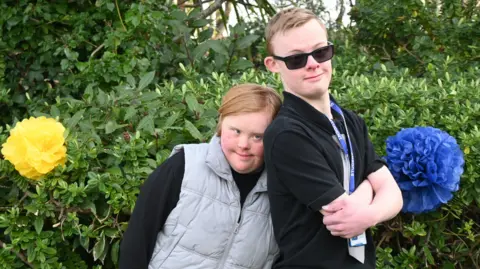 Two students, Immy and Harry, standing together. Immy is resting her head against Harry's shoulder. They are standing in front of a hedge with two large flowers either side of them. One flower is yellow and the other is blue.