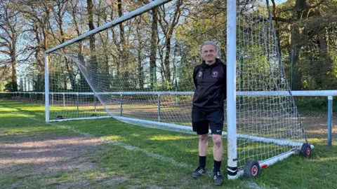 A man leant up against a goal post with his hands behind his back and a Ravenshead Reds jumper on.