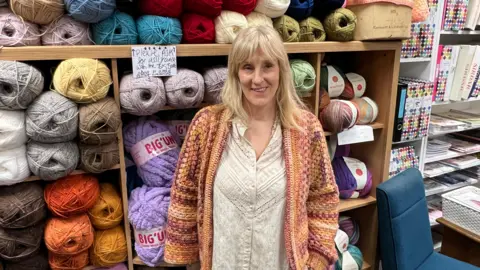 BBC/RICHARD EDWARDS Susie Goate - who runs crafts and fabric shop SewSusieBloom - says she wants the newly elected councillors to build on projects, across Castleford, that are designed to bring the community together, is pictured in her shop. She is standing in front of a rack of huge balls of wool, all different colours. She is smiling at the camera, has shoulder-length blonde hair and is wearing a white blouse and crocheted cardigan.