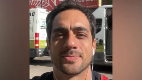 Rodrigo De La Barrera A man with short dark hair and stubble. He is taking a selfie in front of a road.