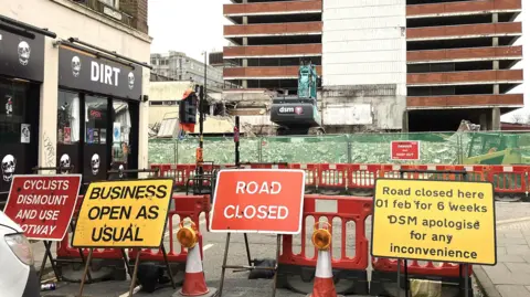 Various road works signs on a road. To the left is a shop called 'Dirt' and in the background is a multi-storey car park.