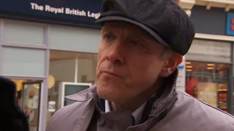 BBC Carl Mould wearing a black cap and grey overcoat