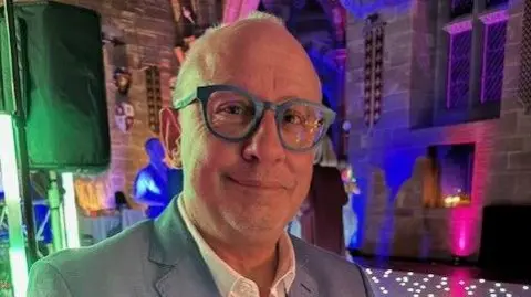 Pete Howell Pete wearing a blue suit with glasses on. There are strobe lights in the background with a dance floor.
