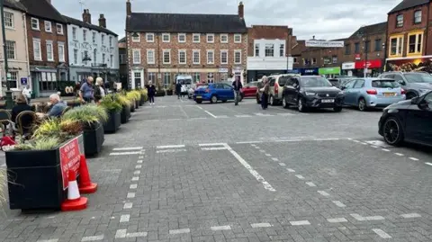 A view of a large market place, laid with grey block paving stones, with parking in the centre and black planters, topped with green shrubs, down one side, separating the road from a pedestrian area. Cars are parked to the right, while people sit at al-fresco dining tables to the left. The square is surrounded by buildings, some Georgian-style, some more modern retail units.