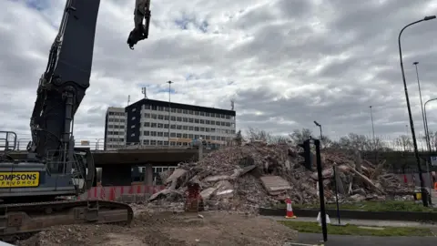 BBC A digger in the foreground on the left-hand side, next to a pile of rubble where Computer House once stood. Behind it is the doomed Gateshead Flyover and another large office block on the other side of the raised road.