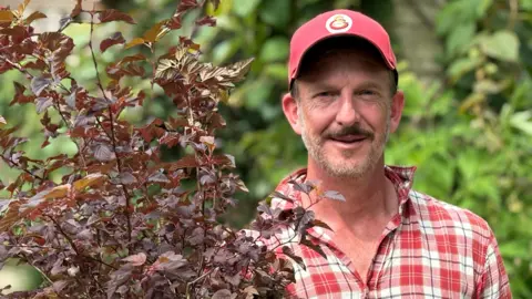 A man in a red and white checked shirt and red baseball cap holds a purple plant. 