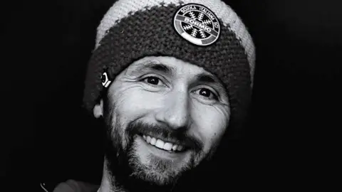 A black and white photo of Justin Loveland. He is smiling broadly, has a beard and is wearing a beanie with Italian words on a badge at the front.