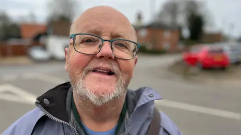 JO THEWLIS/BBC Peter Robertson has short, white hair and a white goatee beard. He is wearing glasses and is looking at the camera.
He is wearing a lilac jacket with a blue t shirt underneath.